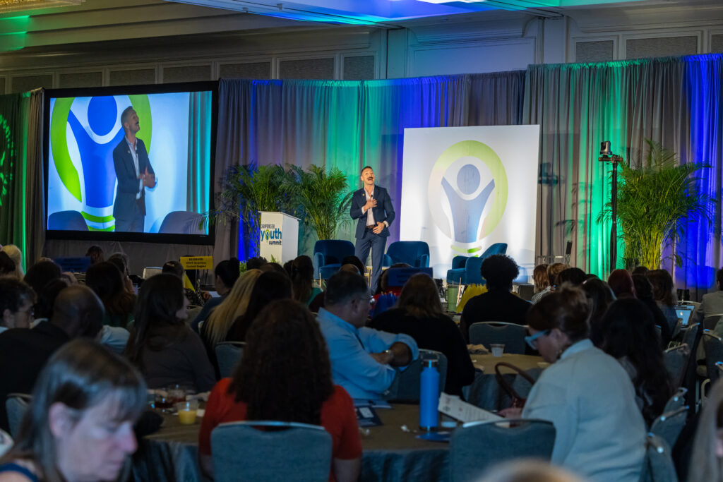 Man on stage speaking to crowd at Champions on Youth Summit 2024.